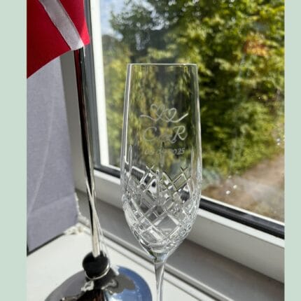 Indgraveret champagneglas ved vindue, dansk flag i baggrund.