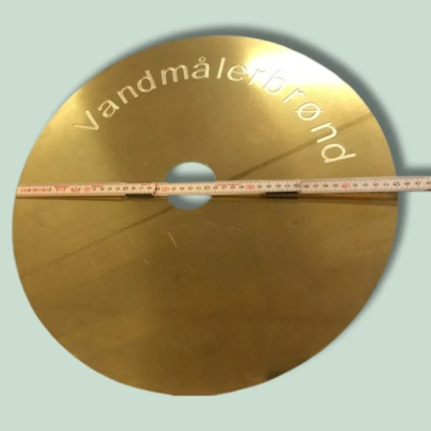 Rund metalplade med teksten "Vandmålerbrønd" og målebånd.